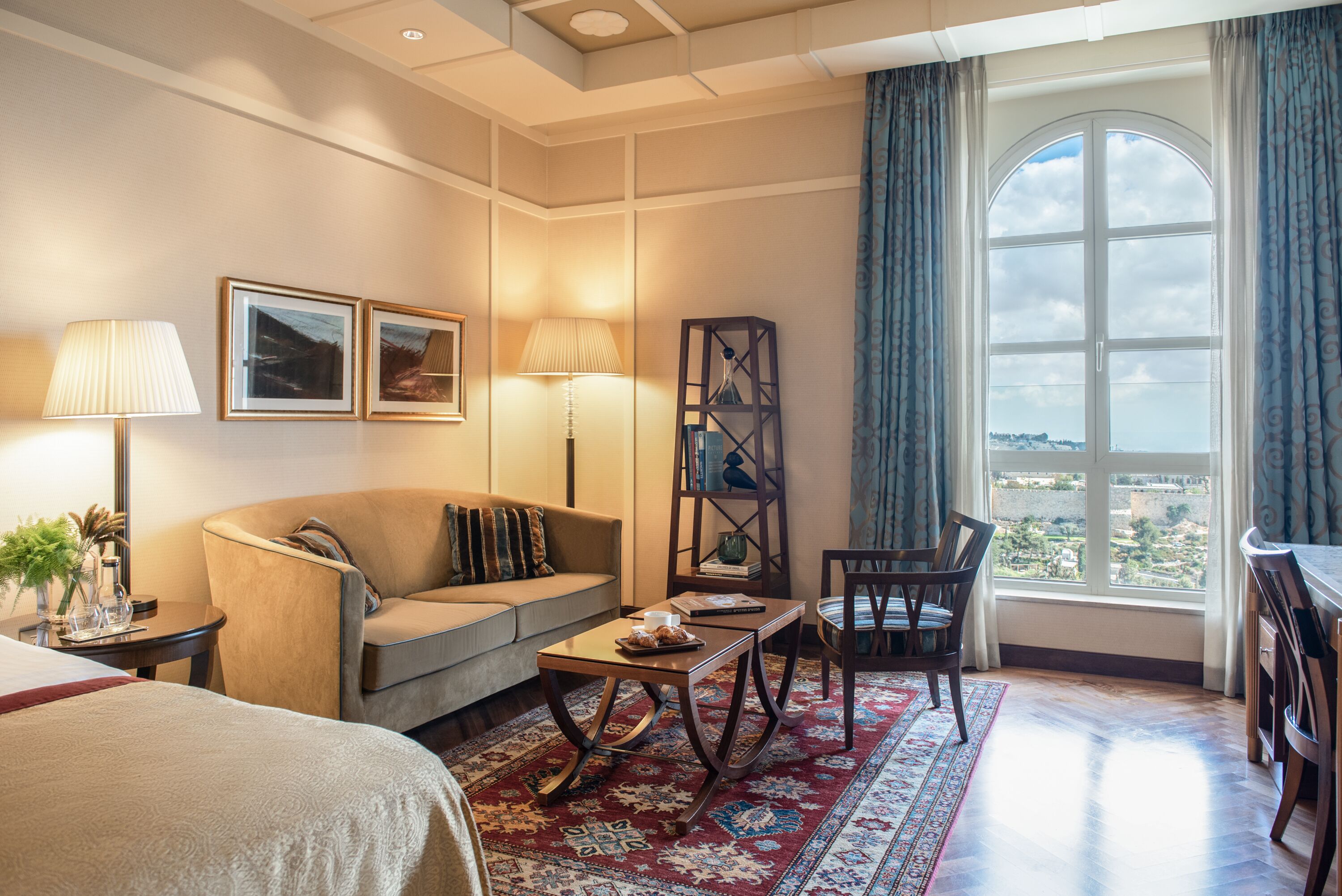 Una habitación de hotel con una cama, un sofá, una mesa de centro, un escritorio, una silla, una lámpara, una estantería y un ventanal con cortinas.