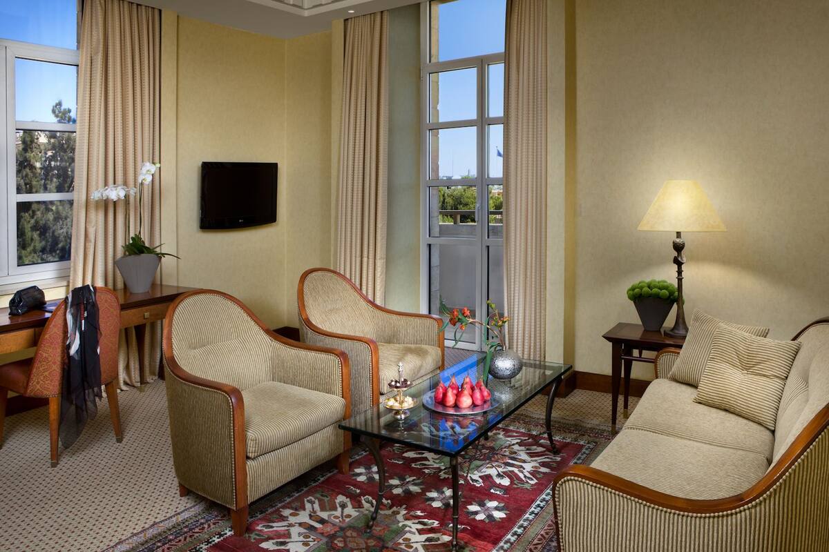 A hotel room with a sofa, two armchairs, a glass coffee table, a TV, a lamp, and a view of the outdoors.