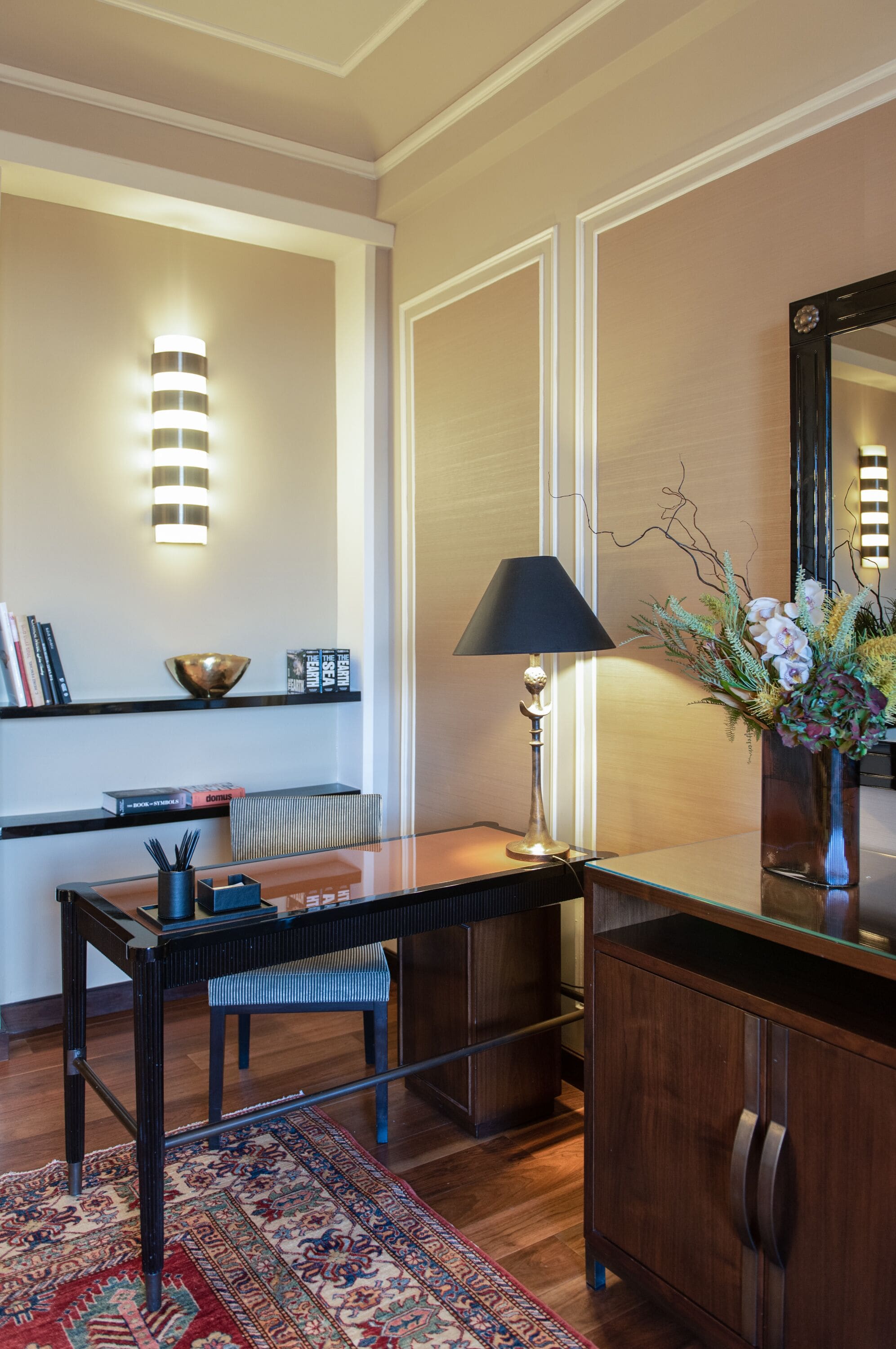 A modern living room with a wooden coffee table, a black lamp, a glass vase with flowers, and a bookshelf with books and a bowl.