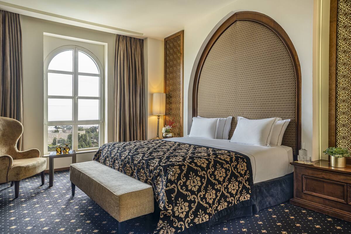 A hotel room with a large bed, a bench, a chair, a window with curtains, and a view of the city.
