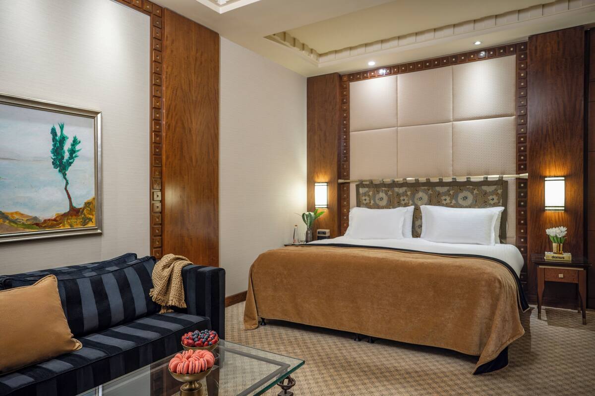 A hotel room with a large bed, a sofa, a glass coffee table with decorative items, and a painting on the wall.
