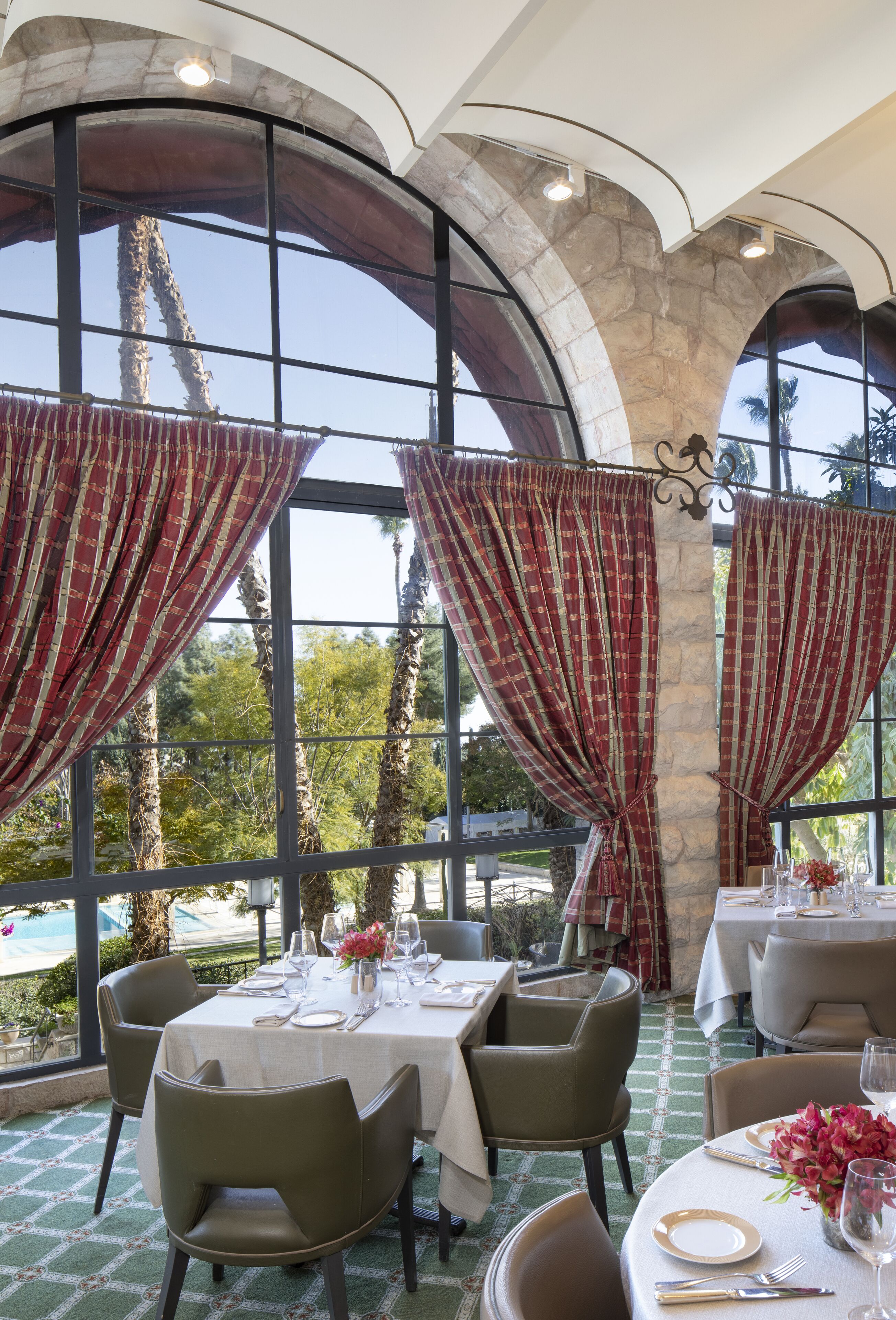 Comedor con mesas dispuestas para dos, ventanas grandes con cortinas y vista a palmeras y una piscina.