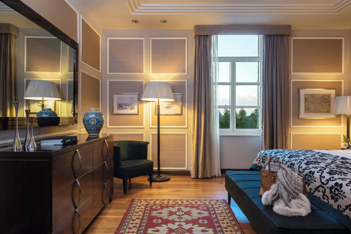 A hotel room with a bed, a dresser, a chair, a floor lamp, and a window with curtains.