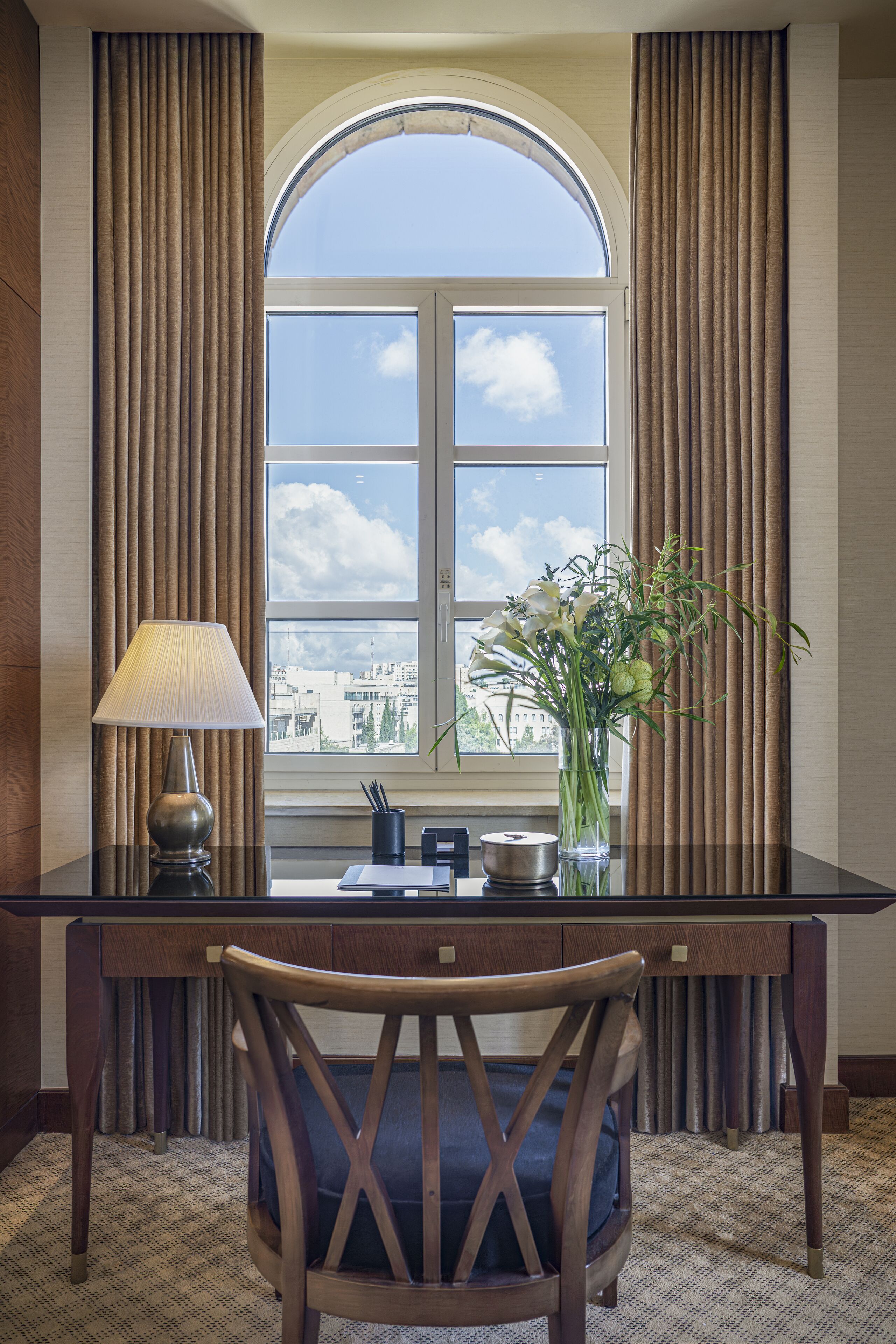 Ein Hotelzimmer mit Schreibtisch, Stuhl, Lampe, einer Vase mit Blumen und einem großen Bogenfenster mit Vorhängen.