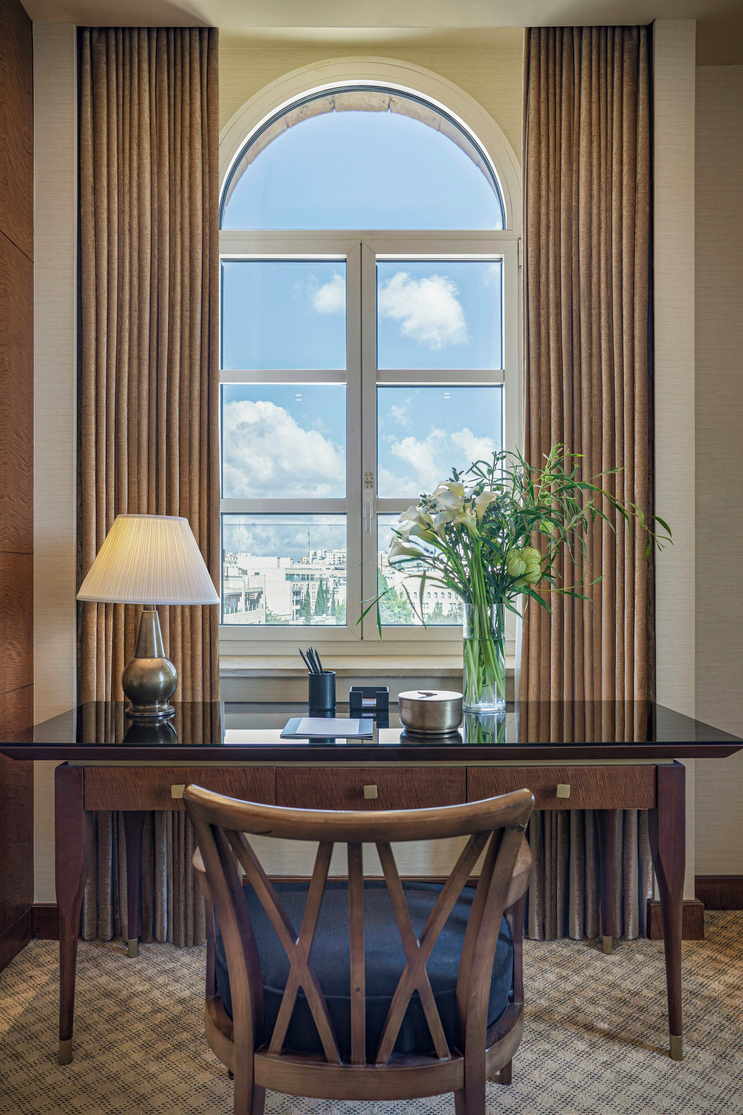 Ett hotellrum med ett skrivbord, en stol, en lampa och utsikt över staden genom ett stort, välvt fönster.