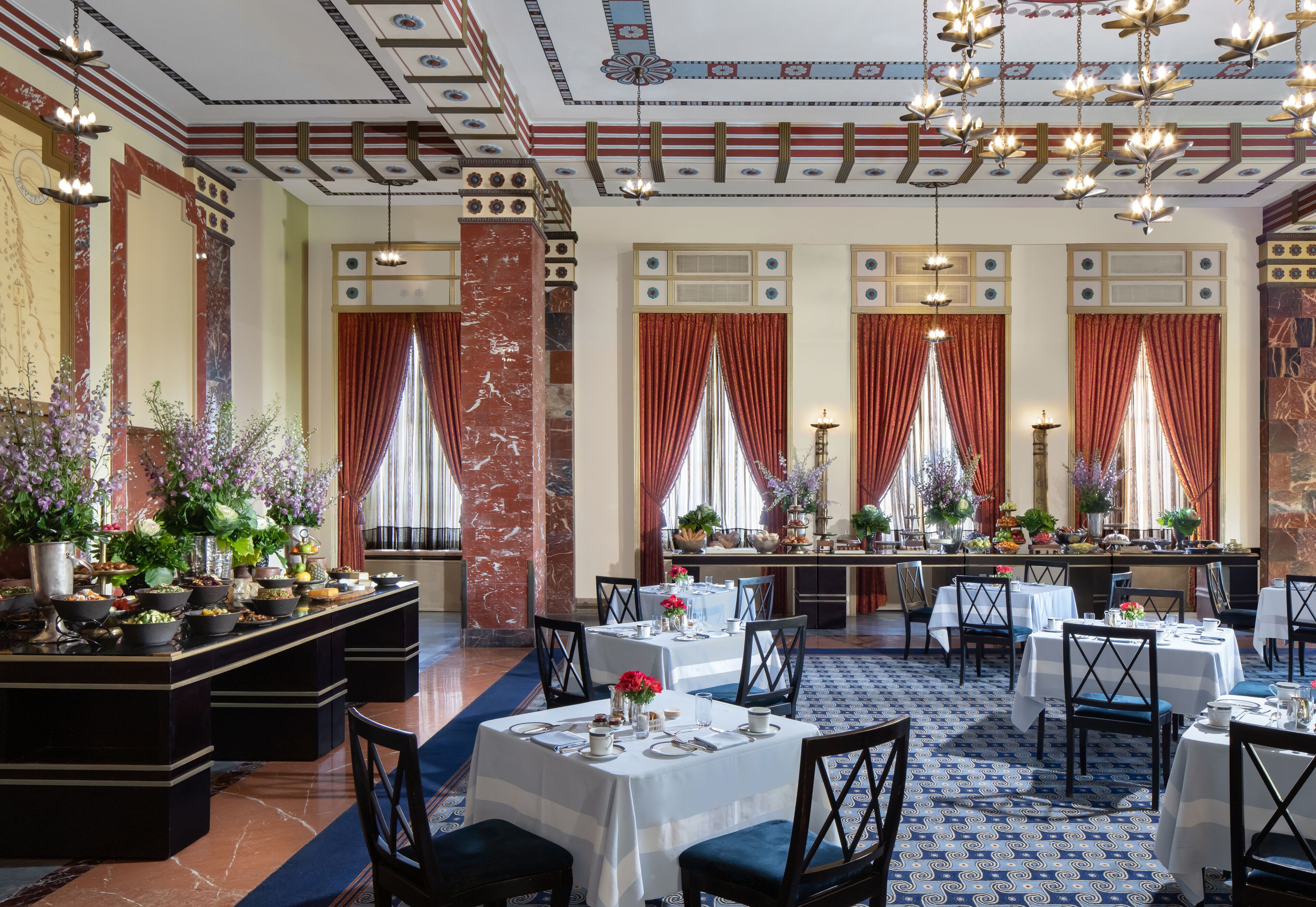Un comedor formal con columnas de mármol, candelabros y cortinas rojas. Las mesas están dispuestas con manteles blancos y sillas.
