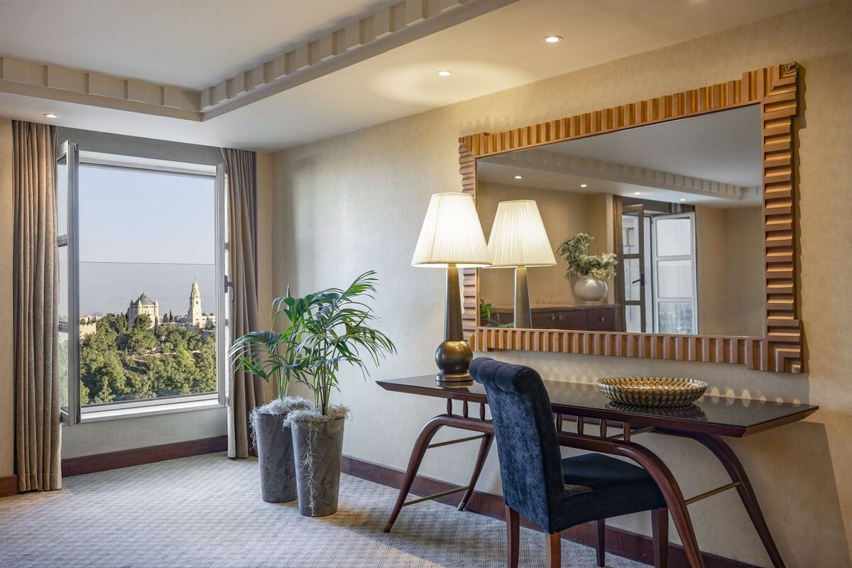A hotel room with a large window, a dining table with a chair, a potted plant, and a mirror with a decorative frame.