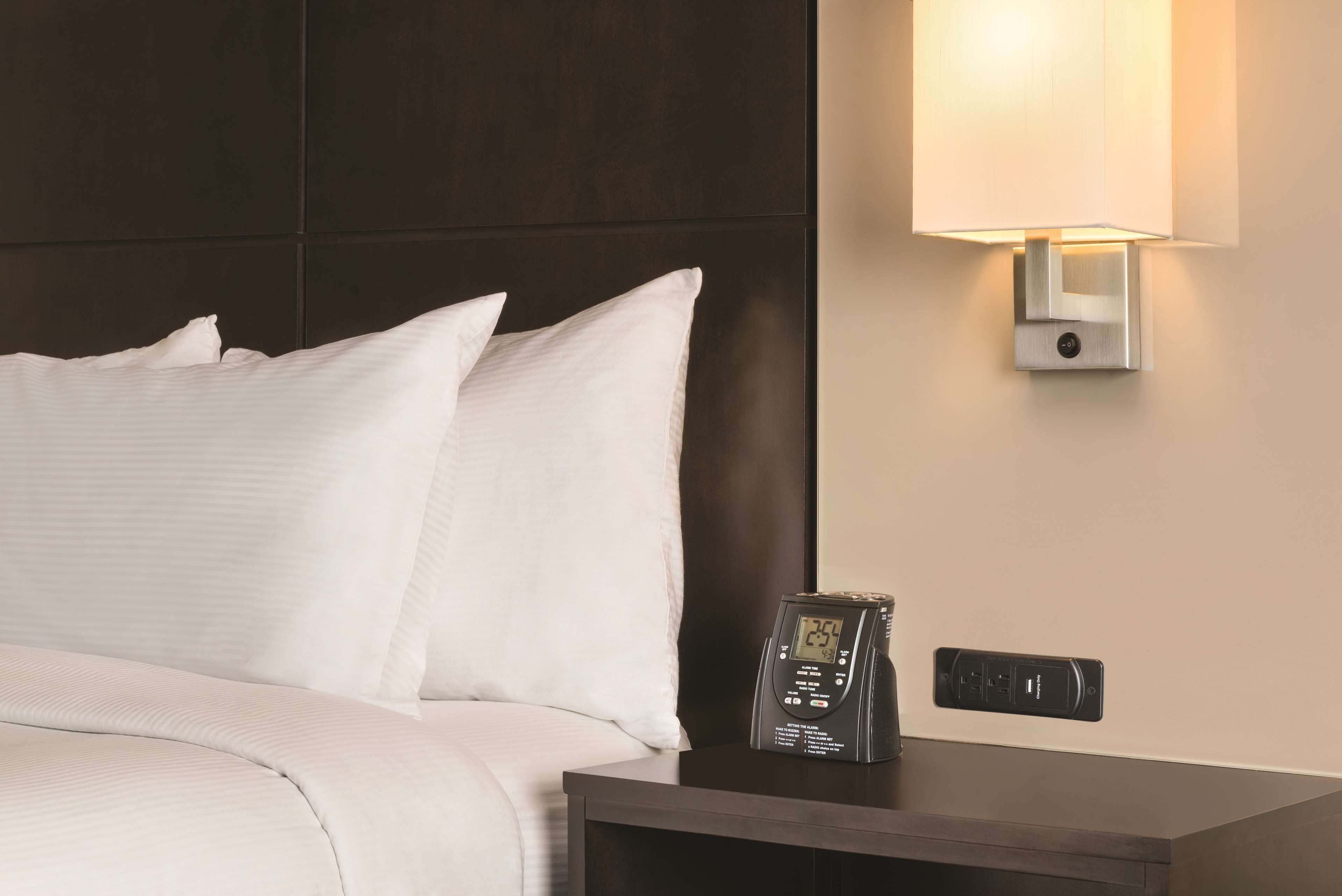 A neatly made bed with white linens, a digital clock on the bedside table, and a wall-mounted lamp.