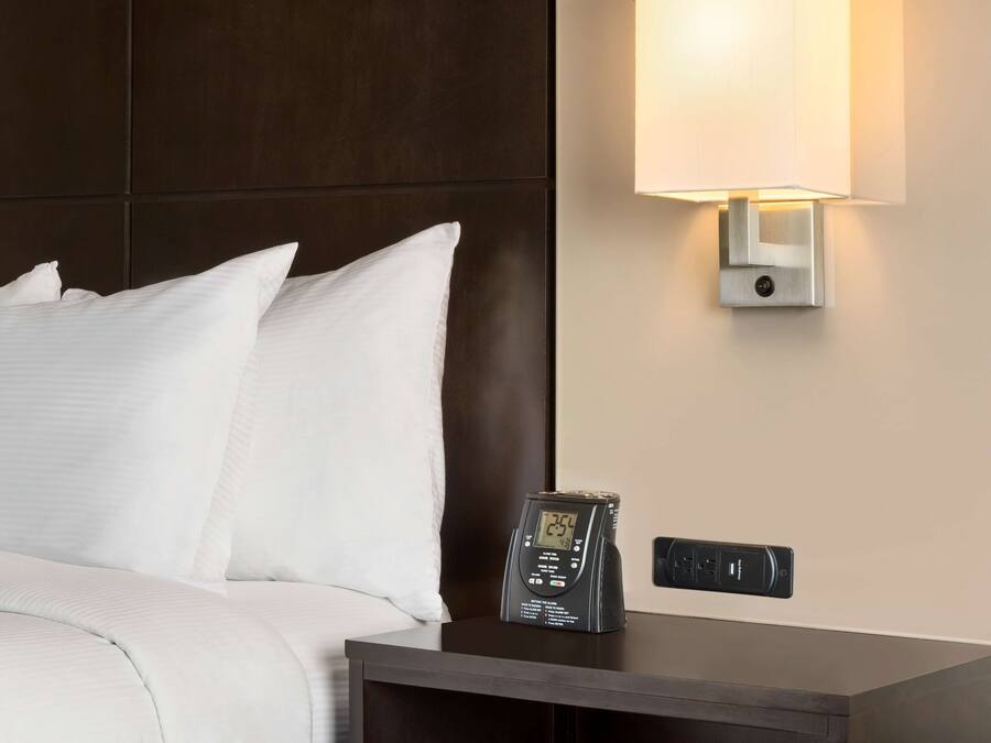 A neatly made bed with white linens, a digital clock on a bedside table, and a wall-mounted lamp.