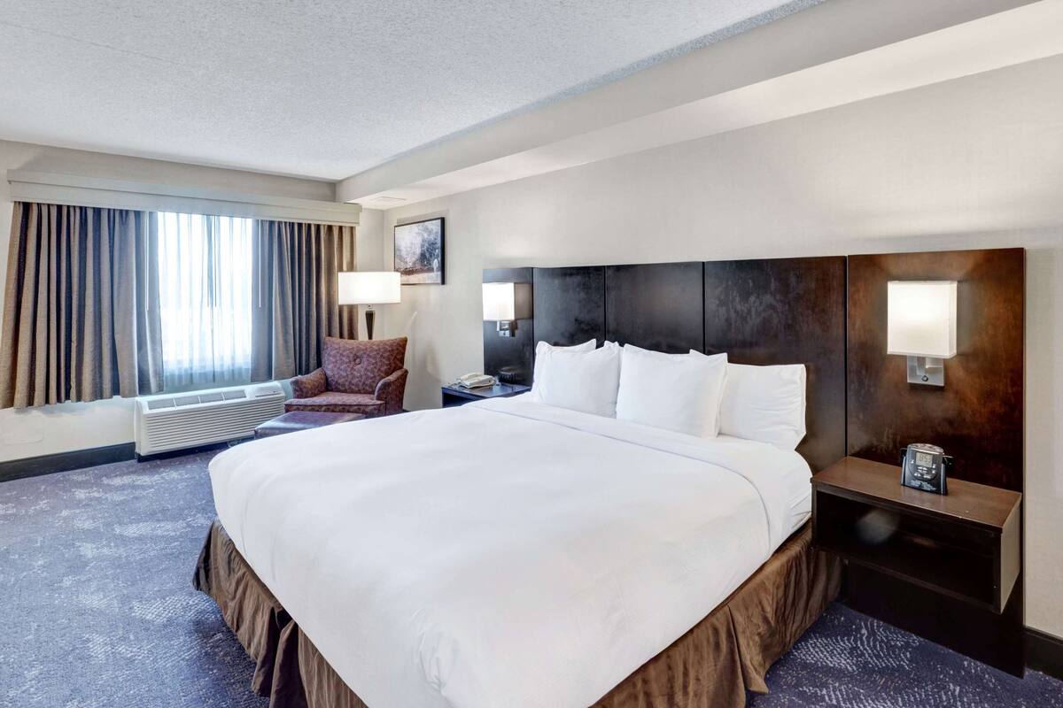 A hotel room with a large bed, a chair, a nightstand, a lamp, and a window with curtains.