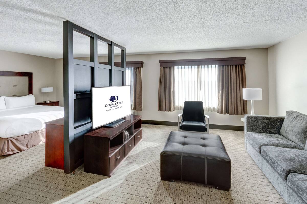 A hotel room with a bed, a TV on a wooden stand, a leather armchair, a grey sofa, and a large window with curtains.