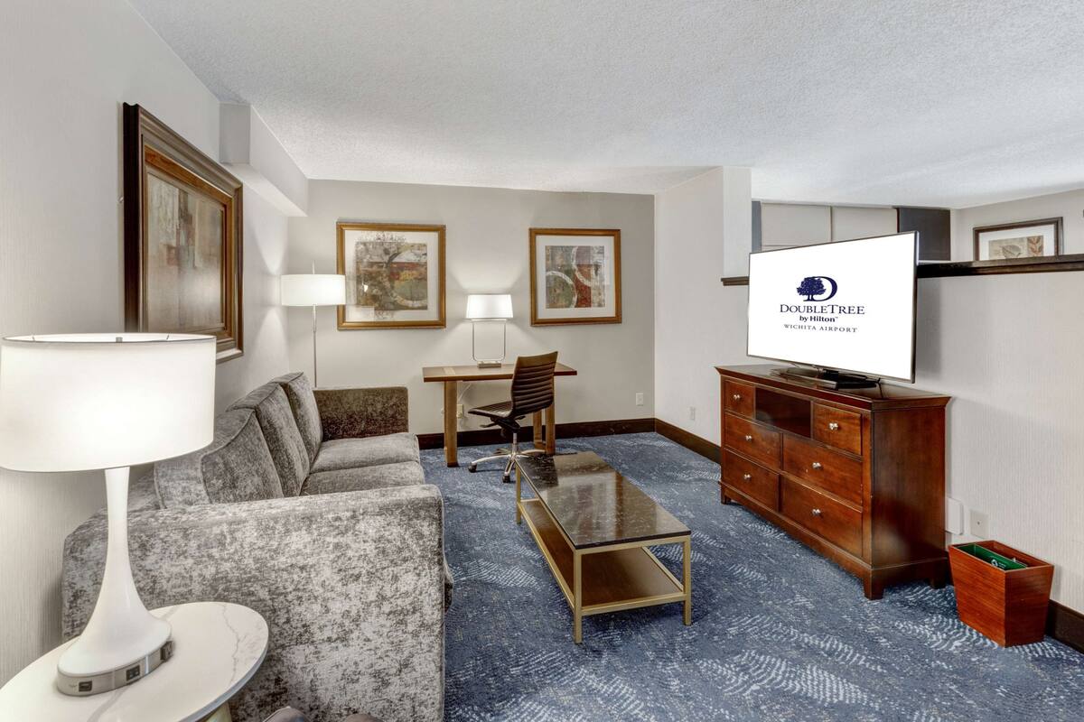 A hotel room with a sofa, armchair, desk, TV, and a small cabinet.