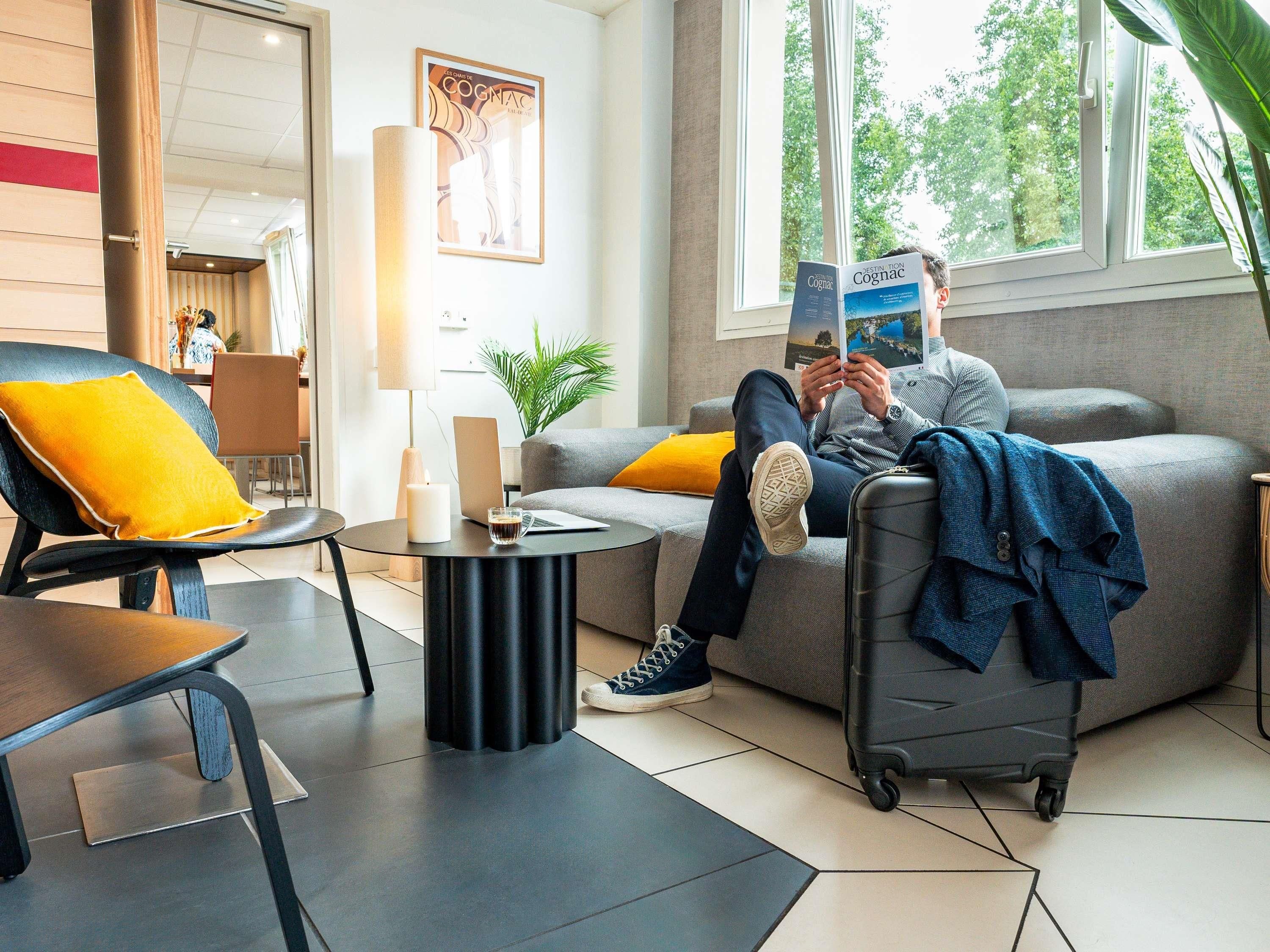 Een moderne hotelkamer met een grijze bank, een zwarte fauteuil en een kleine ronde tafel. Op de bank ligt een laptop en op de vloer een koffer.