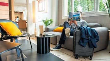Een moderne hotelkamer met een grijze bank, een zwarte fauteuil en een kleine ronde tafel. Op de bank ligt een laptop en op de vloer een koffer.