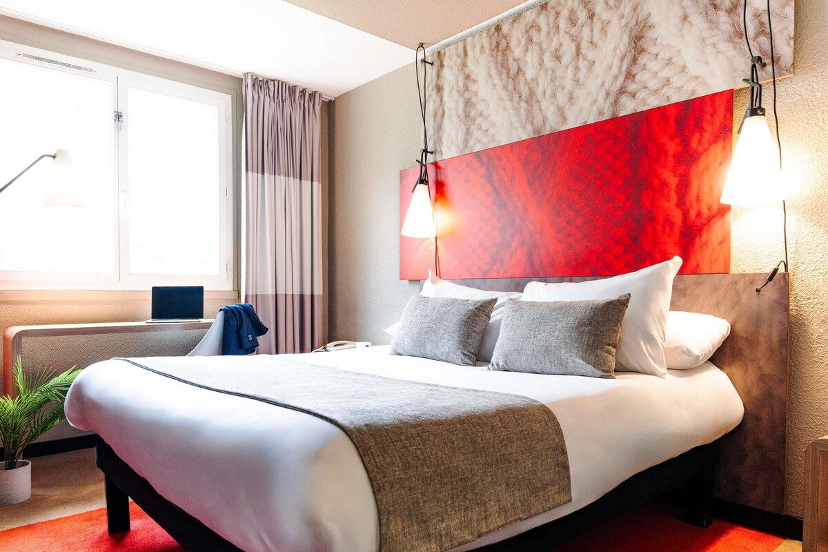A hotel room with a large bed, a bedside table, a lamp, and a view of the outdoors.