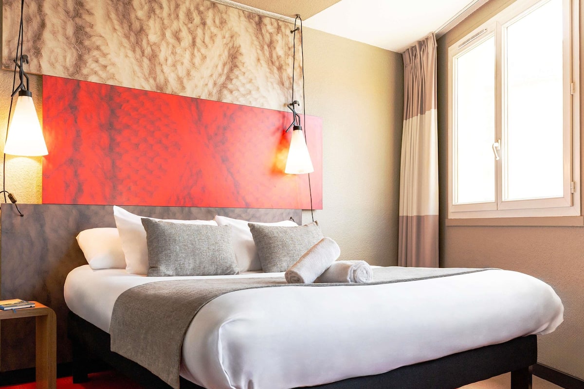 Una habitación de hotel moderna con una cama grande, una pared de acento roja y dos lámparas colgantes.
