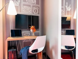 A modern hotel room with a large flat-screen TV, two laptops, a desk with a vase of flowers, and two white chairs.