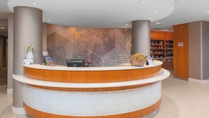 A reception desk with a modern design, featuring a wooden finish and a white countertop. Behind the desk, there is a wall with a geometric pattern and text. - SpringHill Suites Cincinnati North/Forest Park (Cincinnati)