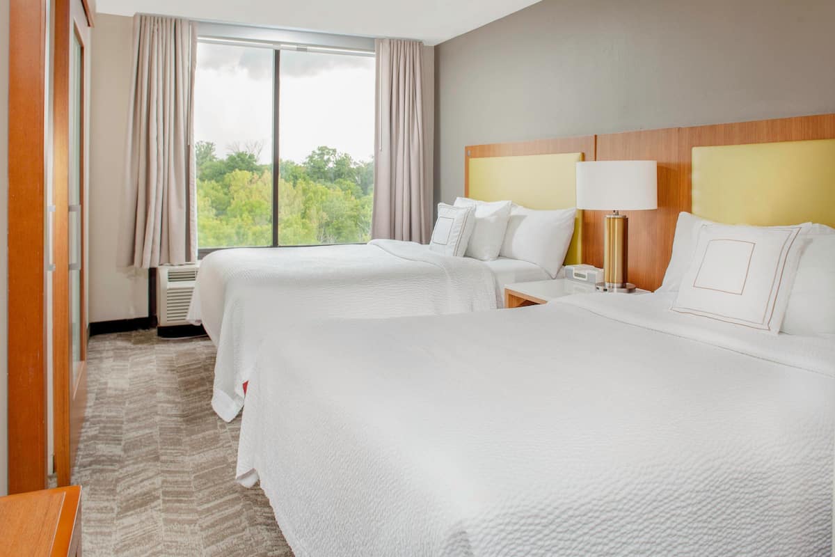 A hotel room with two beds, a wooden headboard, a bedside table with a lamp, and a large window with curtains.