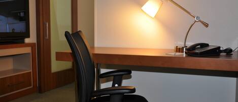A hotel room with a wooden desk, a black office chair, a desk lamp, a telephone, and a television on a stand.