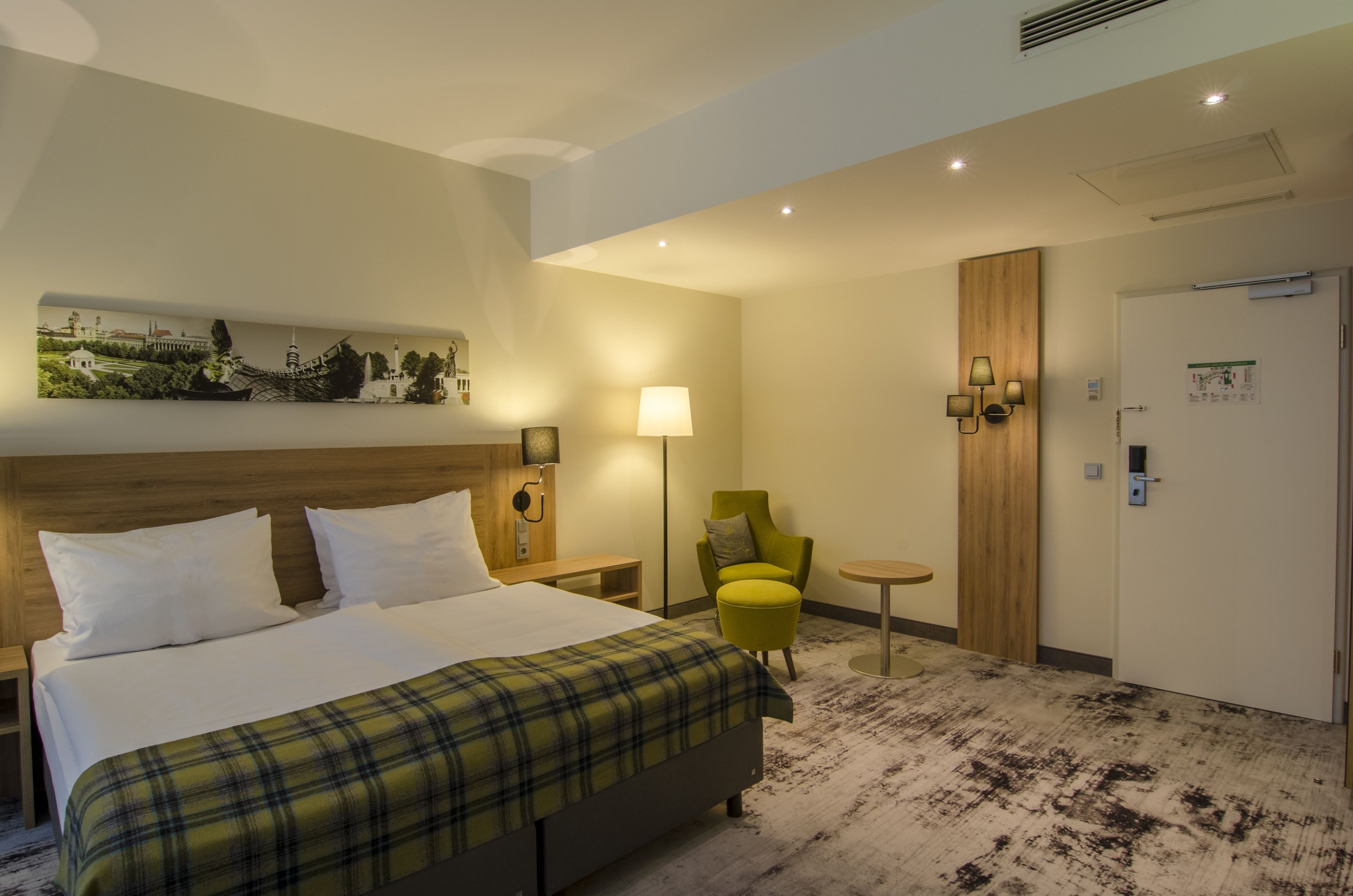 A hotel room with a bed, a green armchair, a small round table, a lamp, and a wall-mounted artwork.