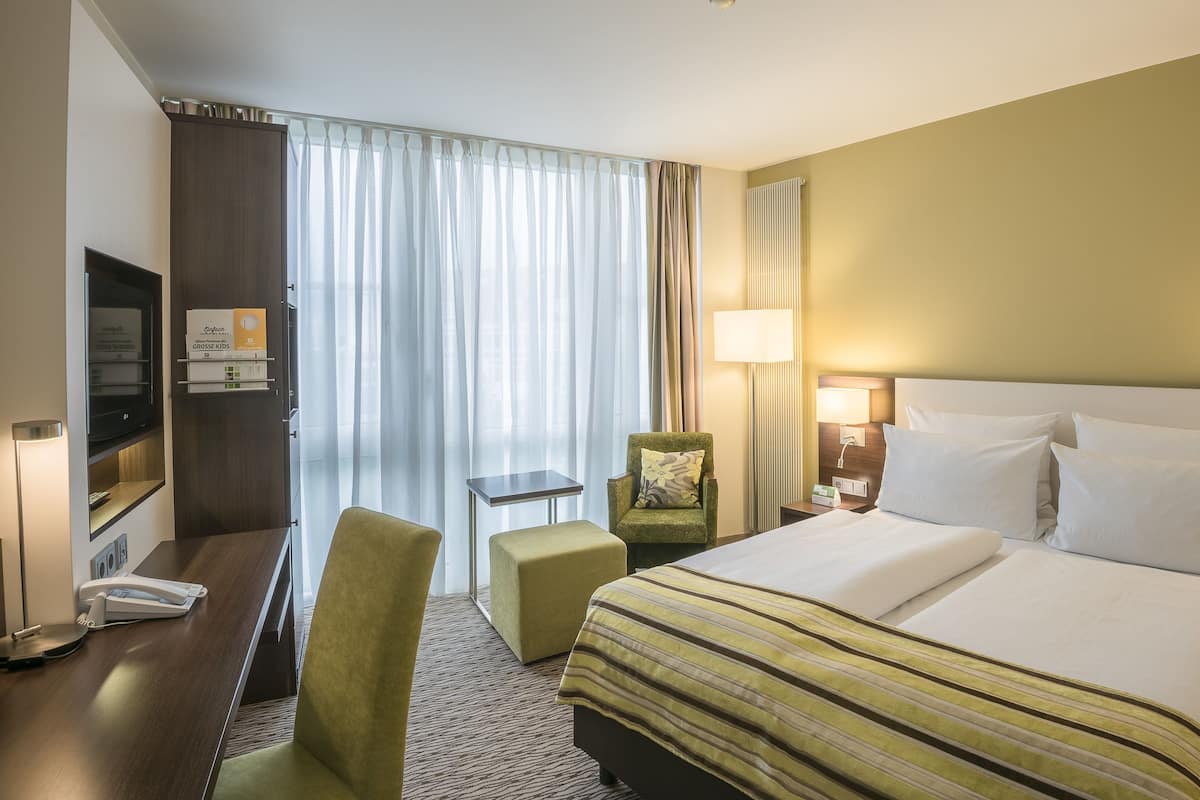 Una camera d'albergo con un letto, una scrivania, una sedia, una televisione e una finestra con tende.