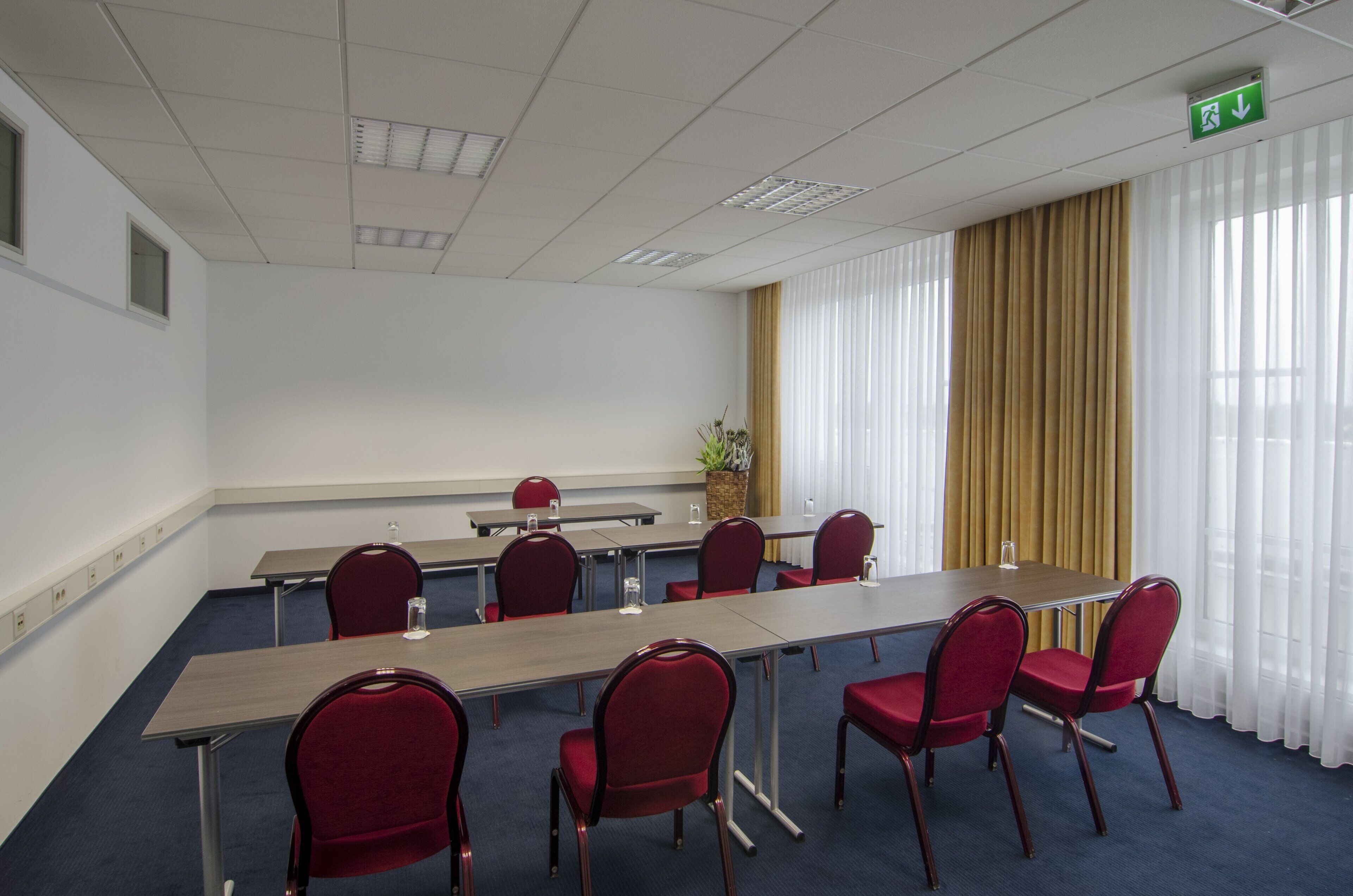 Un salón de conferencias con una larga mesa, sillas rojas, una planta en maceta y un ventanal con cortinas.