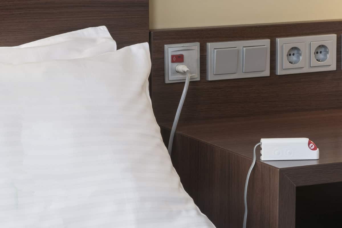 Una camera d'albergo con un letto, un comodino e una presa elettrica con una spina.