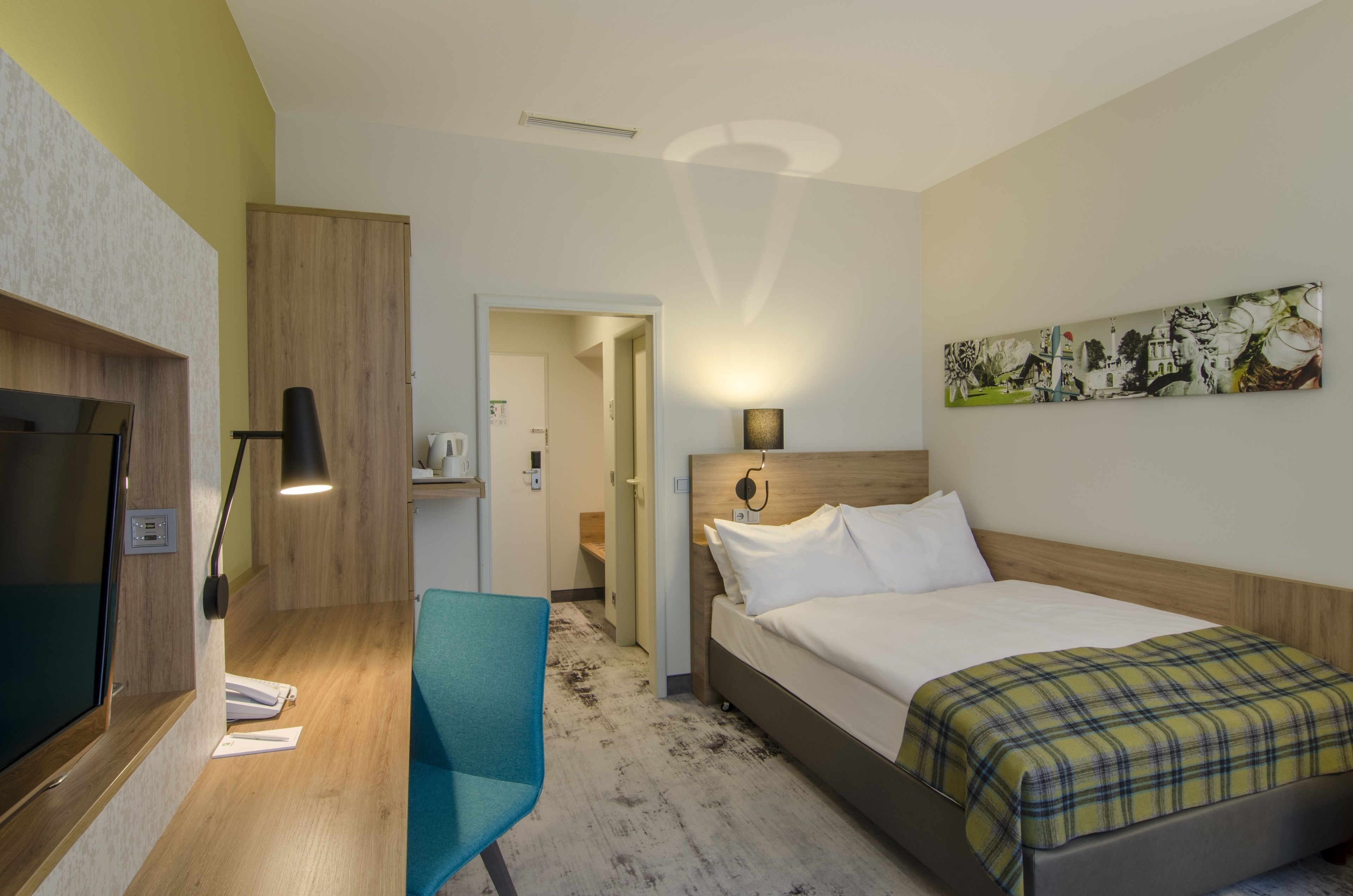 A hotel room with a bed, a desk with a chair, a TV, and a painting on the wall.