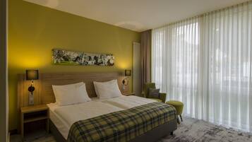 Une chambre d’hôtel avec un grand lit, deux tables de chevet avec des lampes, un fauteuil vert, un tableau au mur et une fenêtre avec des rideaux légers.