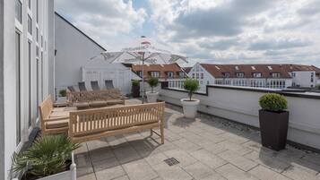 Eine Dachterrasse mit einer Holzbank, Liegestühlen, Blumentöpfen und einem großen Sonnenschirm.