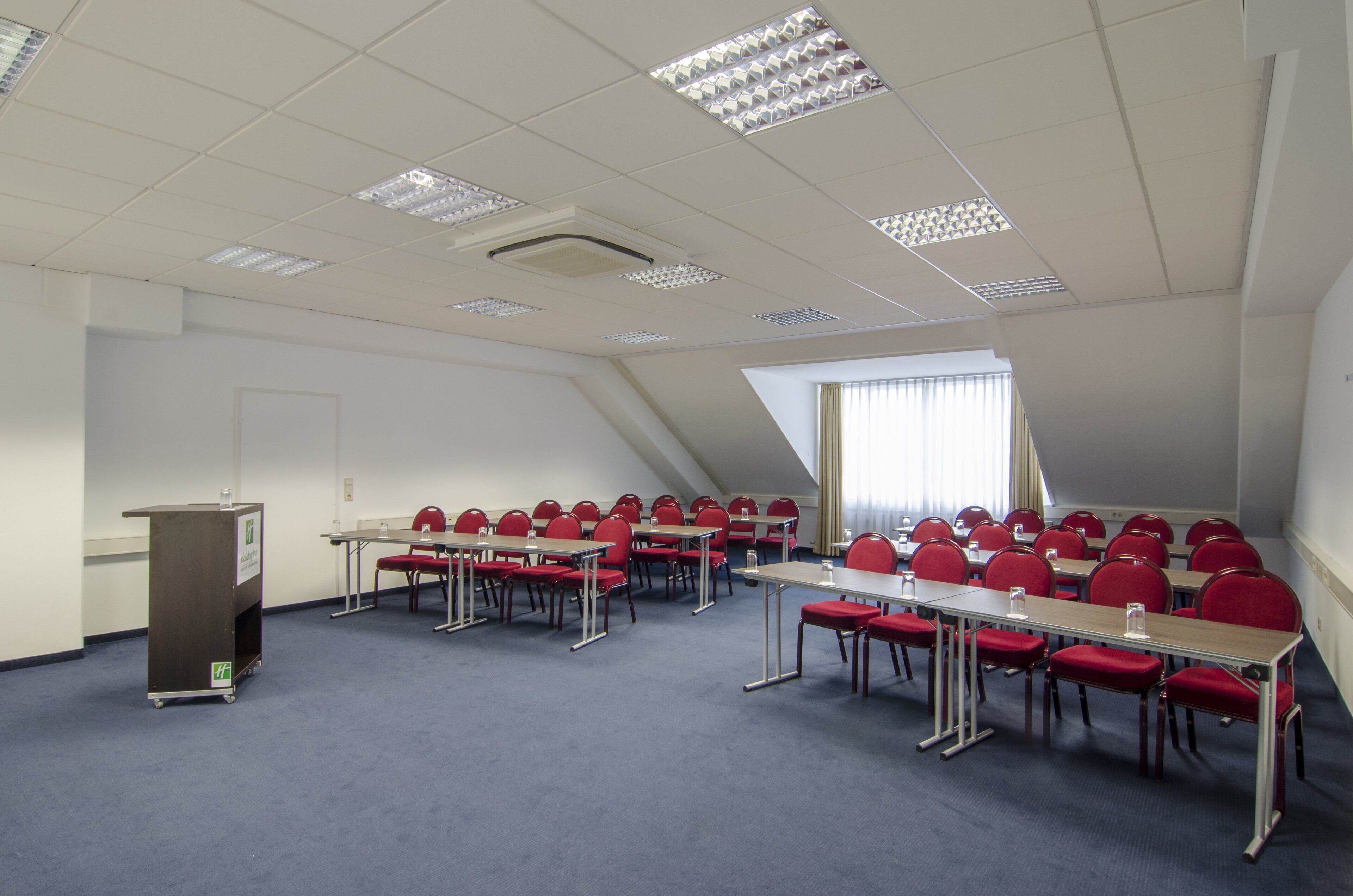 A conference room with red chairs and tables, a podium, and a sloped ceiling with recessed lighting.