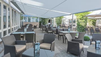 Un patio exterior con mobiliario de mimbre y mesas de vidrio, rodeado por un corredor cubierto y un edificio con grandes ventanales.