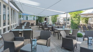 Un patio exterior con mobiliario de mimbre y mesas de vidrio, rodeado por un corredor cubierto y un edificio con grandes ventanales.
