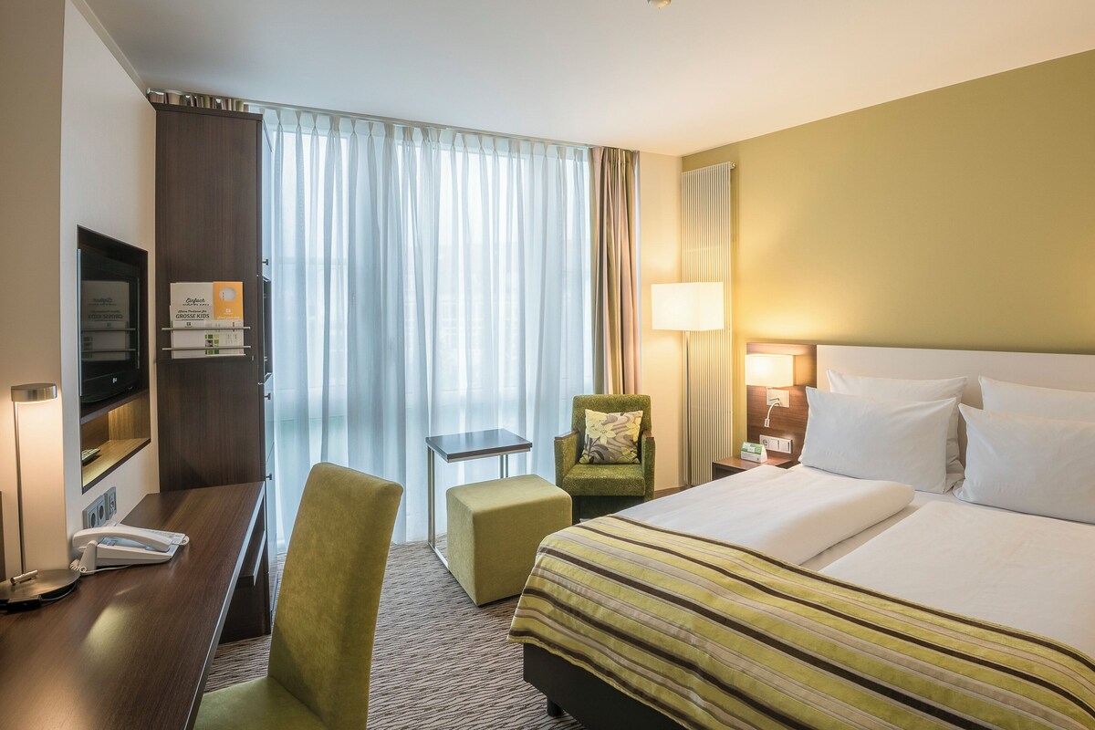 Une chambre d’hôtel avec un lit, un bureau avec un téléphone, une chaise, une lampe et une fenêtre avec des rideaux.