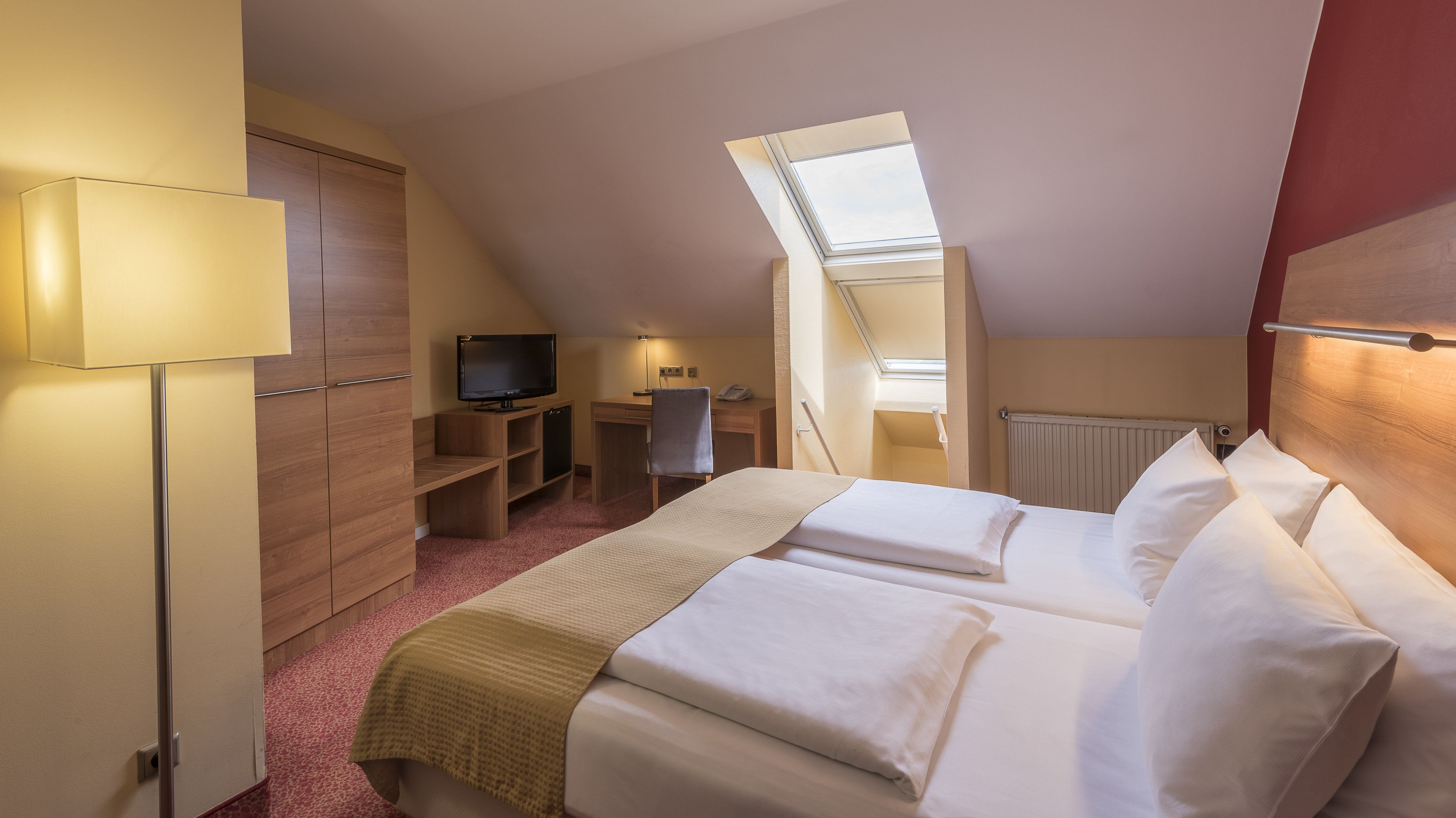 A hotel room with two beds, a desk, a TV, and a skylight.