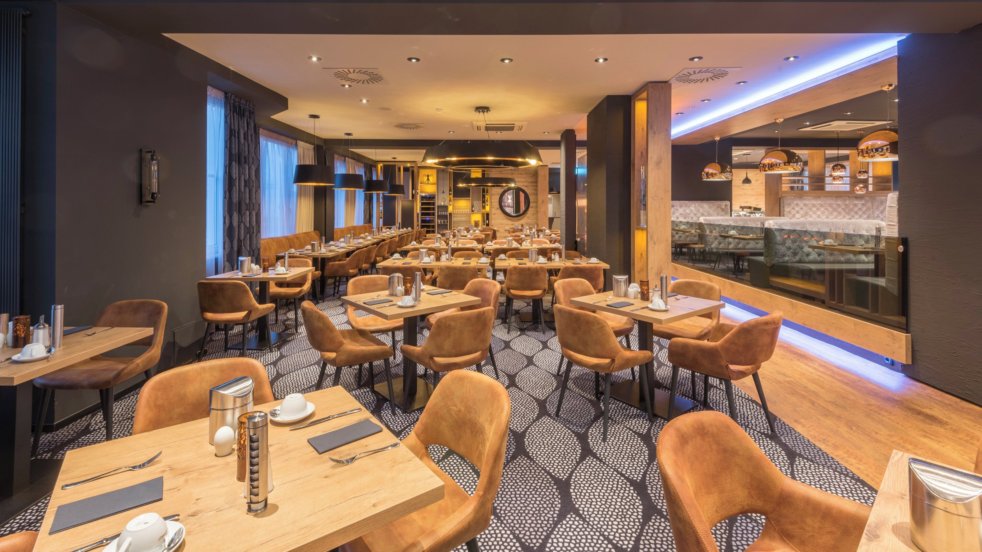 Ein modernes Restaurant mit mehreren Tischen und Stühlen, einem gemusterten Boden und Blick auf den Küchenbereich.