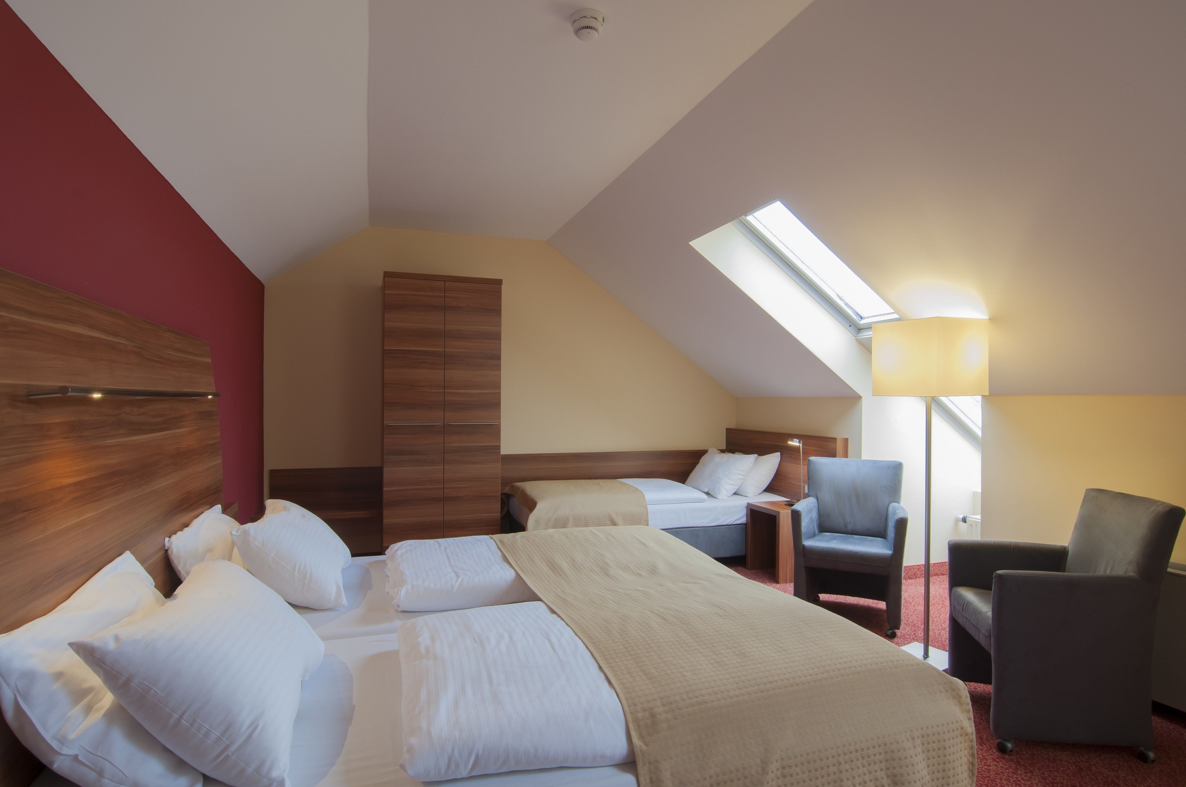 A hotel room with two beds, a red accent wall, a skylight, and a nightstand with a lamp.