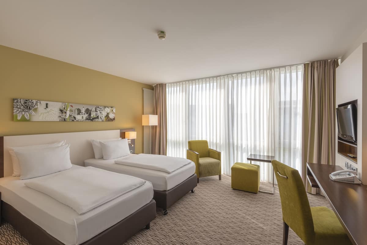 A hotel room with two beds, a desk, a chair, a TV, and a large window with curtains.