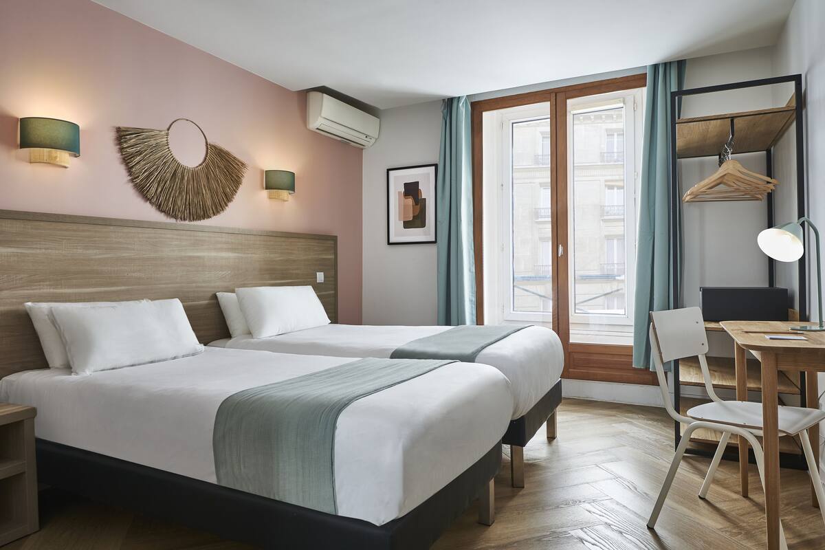 A hotel room with two beds, a desk, a chair, a hanging light fixture, and a wall-mounted air conditioner.