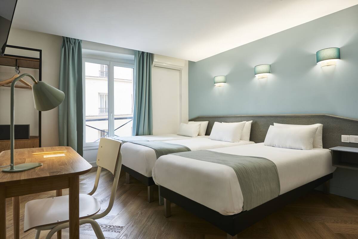 A hotel room with two beds, a desk, and a chair. There are two wall-mounted lamps and a window with curtains.