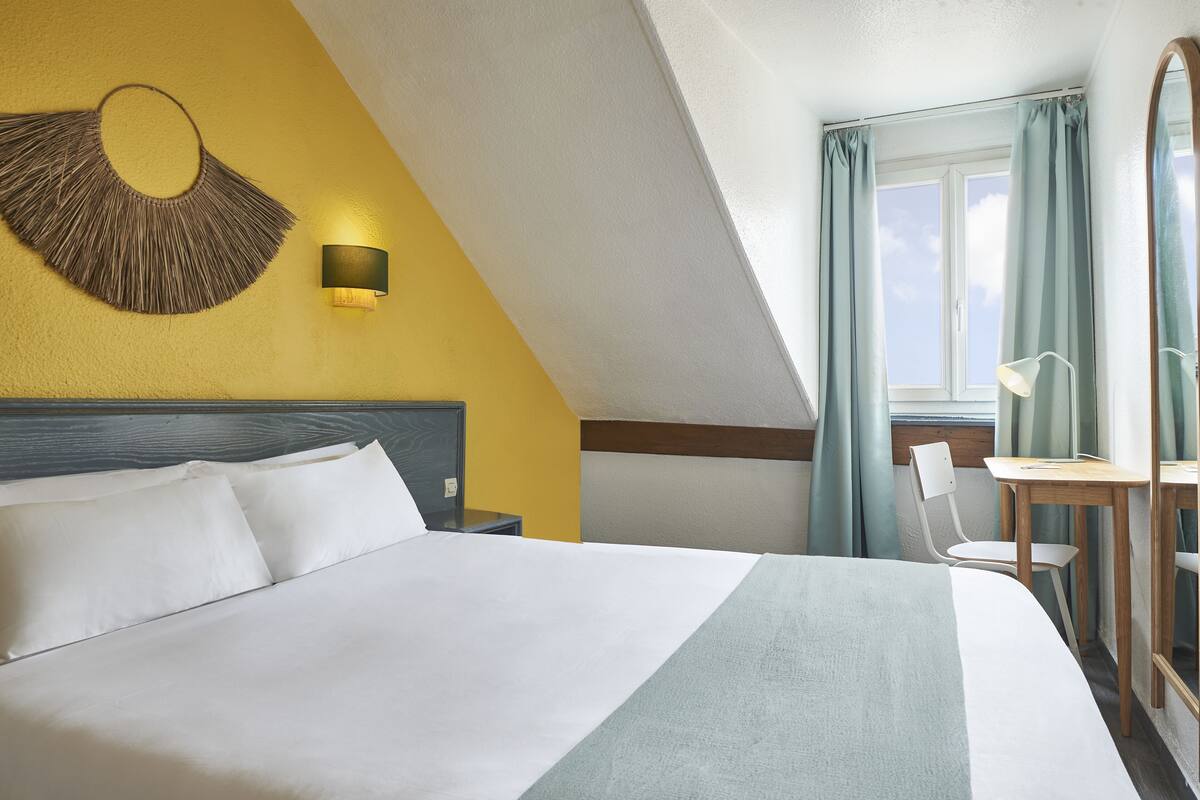 A hotel room with a large bed, a wall-mounted lamp, a desk with a chair, and a window with curtains.