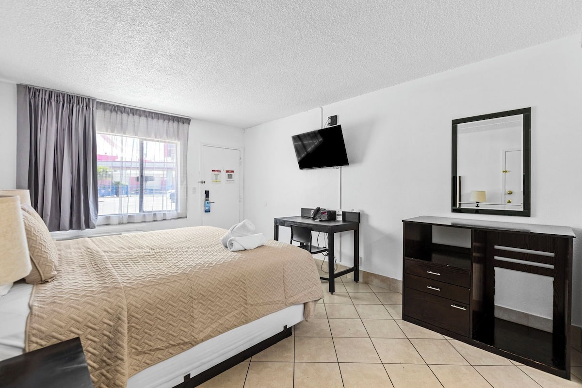A hotel room with a bed, a desk, a flat-screen TV, a mirror, and a window with curtains.