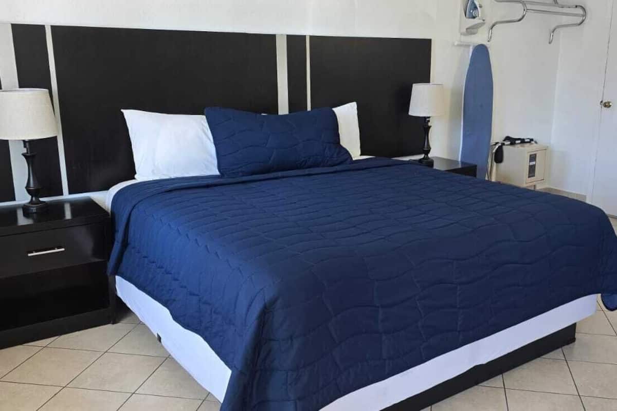 A neatly made bed with a dark blue quilt, two bedside tables with lamps, a small desk with a mirror, and a white wall with a black and white striped panel.