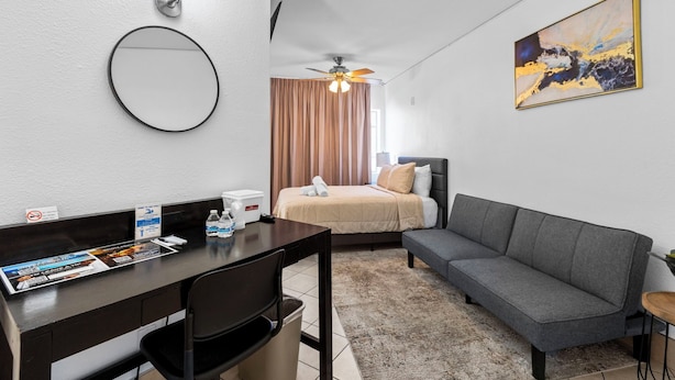 A hotel room with a bed, a sofa, a desk with a chair, a ceiling fan, a wall-mounted light, and a painting.