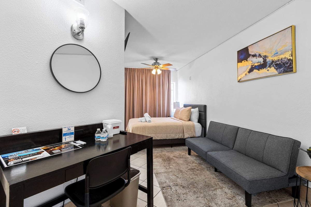 A hotel room with a bed, a sofa, a desk with a chair, a ceiling fan, a wall-mounted light, and a painting.