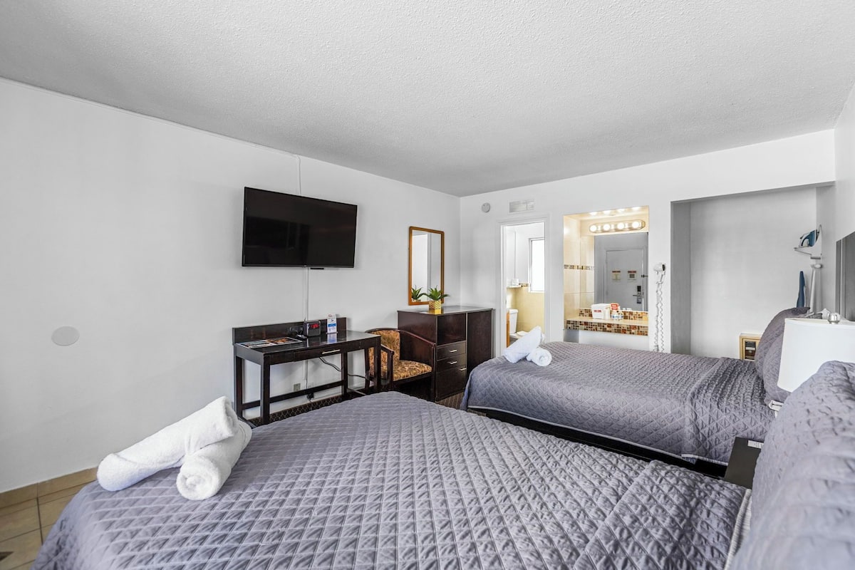 A hotel room with two beds, a desk, a TV, a mirror, and a bathroom with a vanity and mirror.