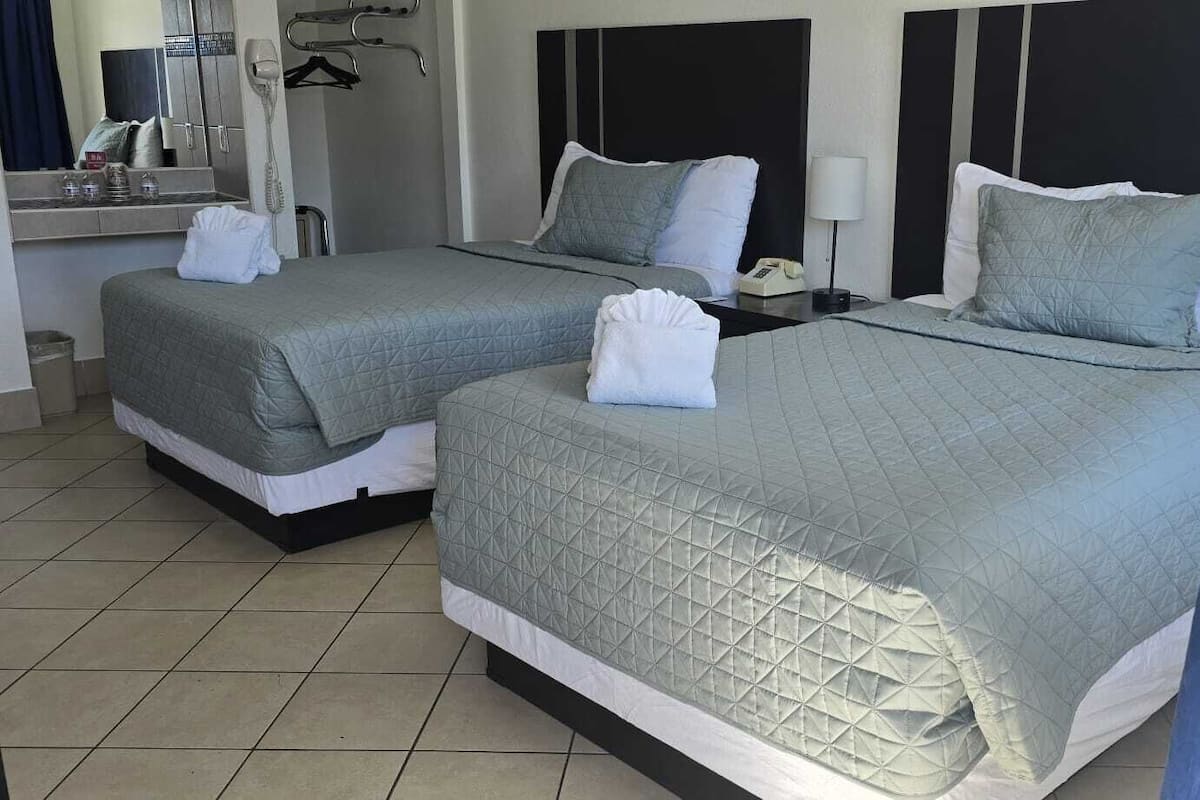 A hotel room with two beds, each with a quilted bedspread and pillows, a bedside table with a lamp, and a mirror above the bed.