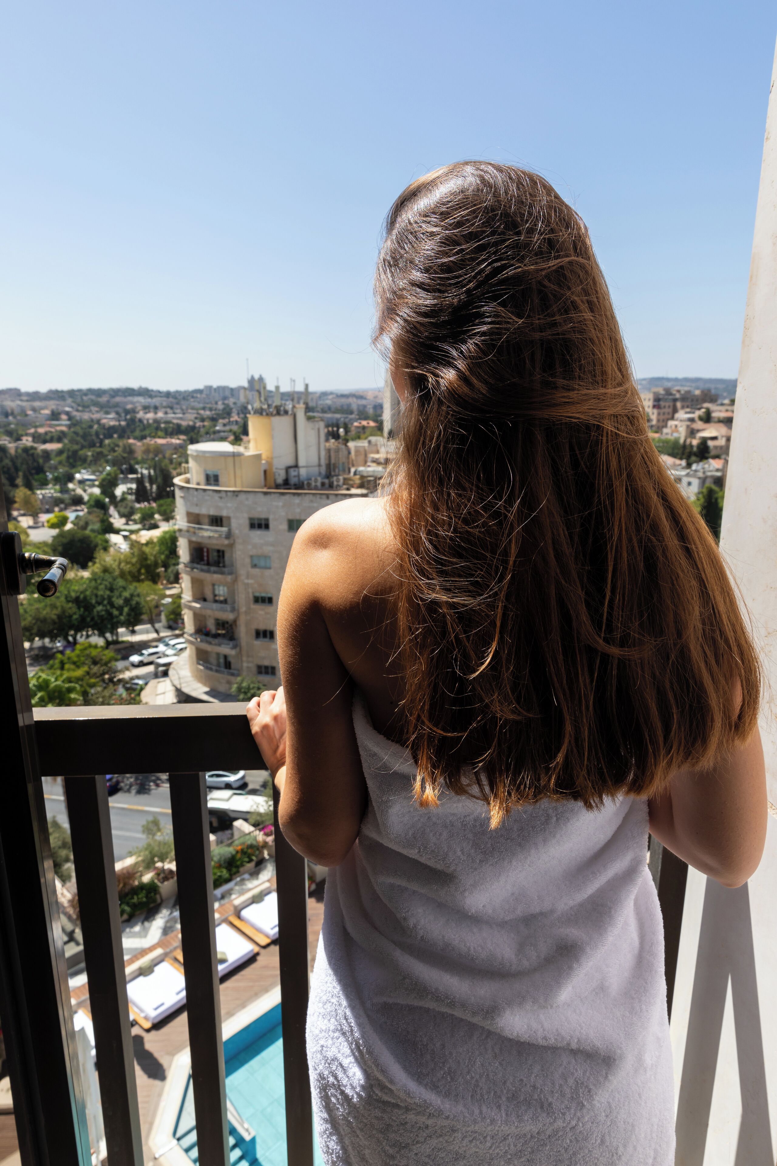 A person standing on a balcony overlooking a cityscape with buildings and a pool area.