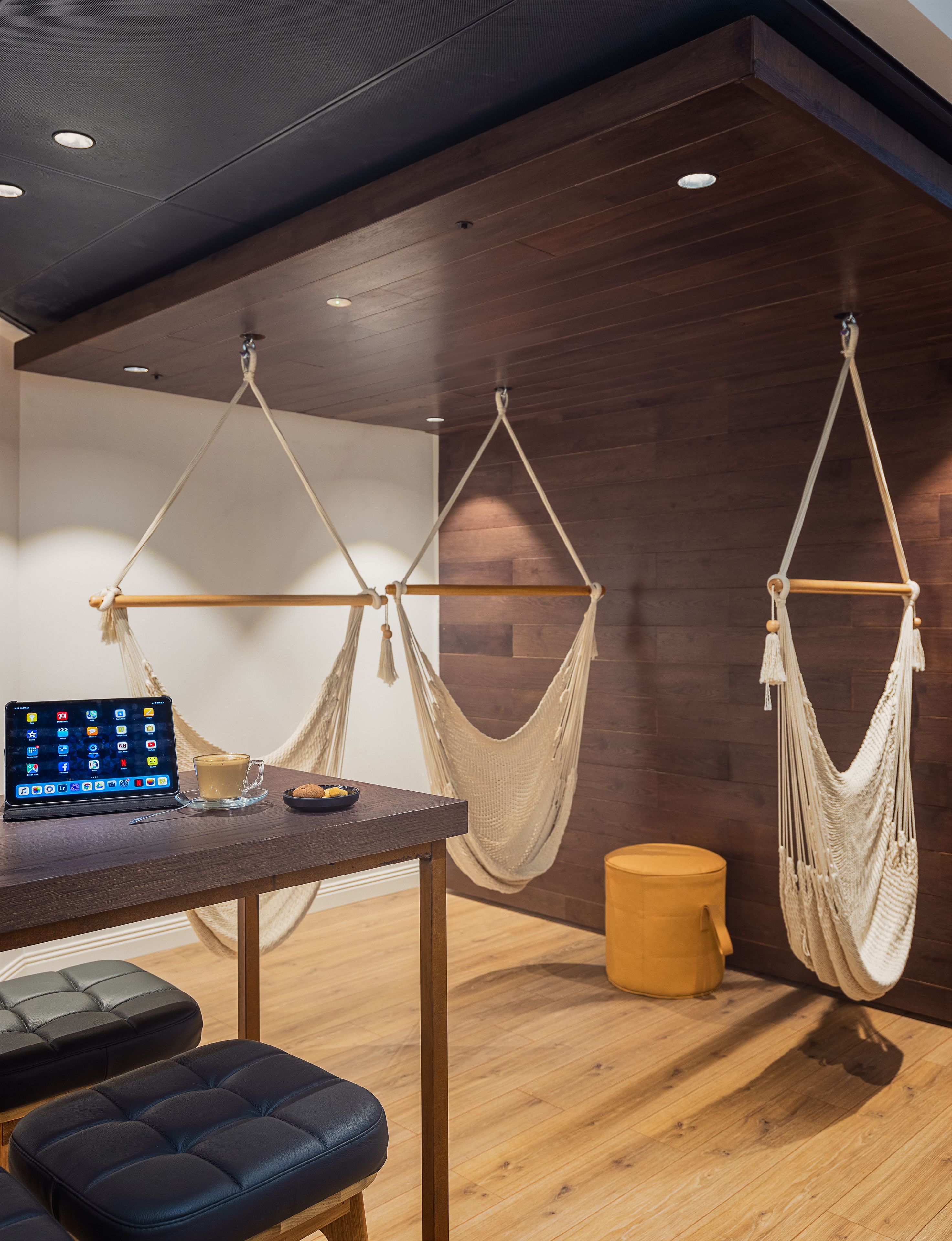 A modern room with a wooden floor, a desk with a tablet and coffee cup, two hammocks, and a yellow ottoman.